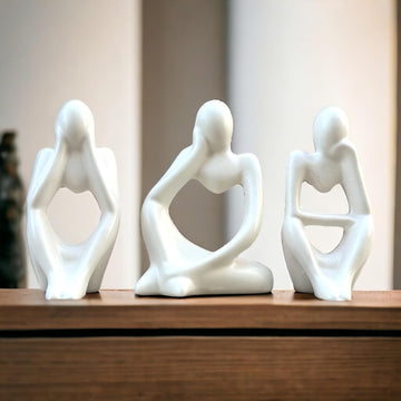 Three white ceramic sculptures of people sitting on a wooden surface with a neutral background