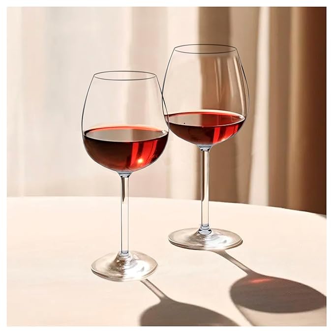 Two clear borosilicate wine glasses on a table, filled with red wine, with a window in the background allowing natural light to come in.