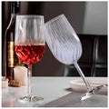 Two clear borosilicate red wine glasses, one filled with red wine, displayed on a table with a bottle and other dining accessories in the background.