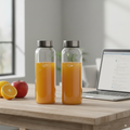 Two glass bottles with orange juice on a desk next to a laptop and fruits.